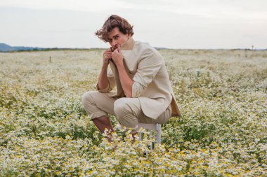 Beyaz takım elbiseli uzun boylu, yakışıklı bir adam papatya çiçekleri tarlasında bir sandalyenin arkasında oturuyordu.