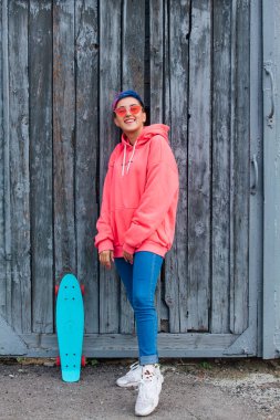 Kısa saçlı, pembe kapüşonlu ve pembe gözlüklü şık bir genç kadın. Plastik kaykayıyla eski ahşap kapıların yanında duruyor. Gençlik kavramı.