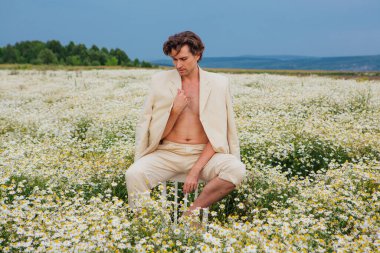 Beyaz takım elbiseli uzun boylu, yakışıklı bir adam papatya çiçekleri tarlasında bir sandalyenin arkasında oturuyordu.