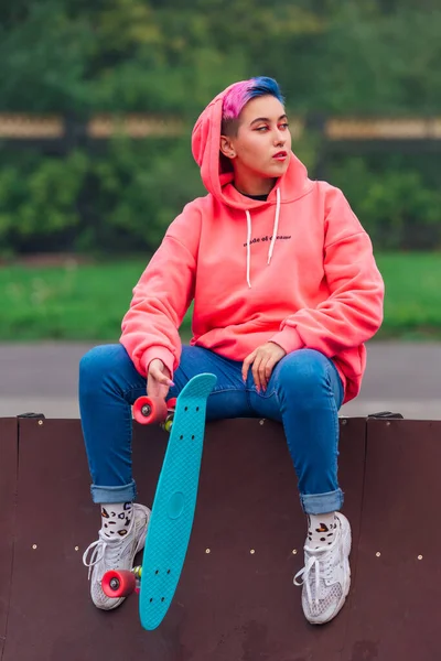 Portrait of a trendy pretty young girl sitting next to the skateboard court with her plastic skateboard. — Stock Photo, Image