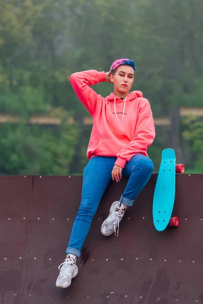 Portret van een trendy mooie jonge gir met kort gekleurd haar naast het skateboard court met haar plastic skateboard. — Stockfoto