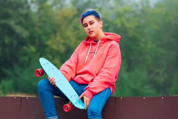 Zomer levensstijl beeld van trendy mooie jonge meisje met kort gekleurd haar zitten naast de skateboard court met haar plastic skateboard. — Stockfoto