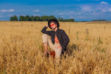 Uzun boylu, siyah şapkalı, pembe gözlüklü, altın yulaf tarlasında kahverengi deri bir valizde oturan yakışıklı bir adam..