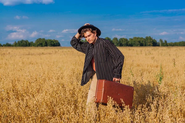 Tall handsome man with black hat and brown suitcase standing at golden oat field.