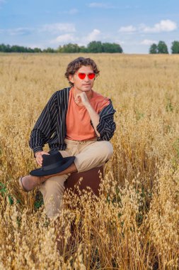 Kırsal Kır Sahnesi. Siyah gömlekli, siyah şapkalı ve pembe gözlükle altın yulaf tarlasında kahverengi deri bir valizde oturan uzun boylu yakışıklı bir adam. Mavi gökyüzü ile yaz manzarası