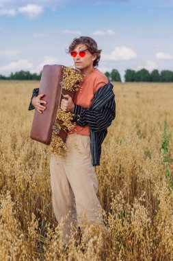 Kırsal Kır Sahnesi. Siyah gömlek ve pembe güneş gözlüğü takmış uzun boylu yakışıklı bir adam altın yulaf tarlasında duruyor. Kahverengi, eski, deri, içi yulaf dolu bir valizle. Mavi gökyüzü ile yaz manzarası