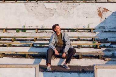 Gün batımında eski stadyumun ezilmiş tribününde kot ceket giymiş uzun boylu yakışıklı bir adamın moda portresi.