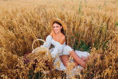 Kırsal Kır Sahnesi. Uzun saçlı, beyaz bluz ve hasır şapkalı güzel bir kadın altın yulaf tarlasında, sepetin yanında, yulaf kulaklı oturuyor..