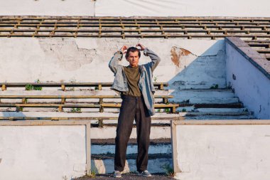 Gün batımında eski stadyumun ezilmiş tribününde kot ceket giymiş uzun boylu yakışıklı bir adamın moda portresi.