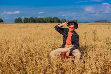 Kırsal Kır Sahnesi. Siyah gömlekli, siyah şapkalı ve pembe gözlükle altın yulaf tarlasında kahverengi deri bir valizde oturan uzun boylu yakışıklı bir adam. Mavi gökyüzü ile yaz manzarası