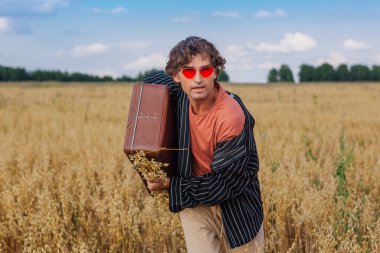 Kırsal Kır Sahnesi. Siyah gömlek ve pembe güneş gözlüğü takmış uzun boylu yakışıklı bir adam altın yulaf tarlasında duruyor. Kahverengi, eski, deri, içi yulaf dolu bir valizle. Mavi gökyüzü ile yaz manzarası