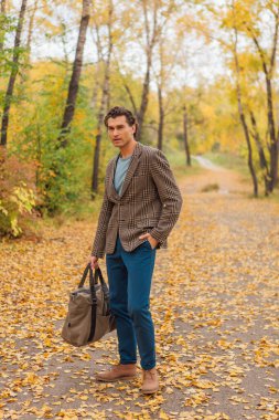 Kahverengi ceketli uzun boylu yakışıklı bir adam sonbahar sokağında çantasıyla yürüyor.