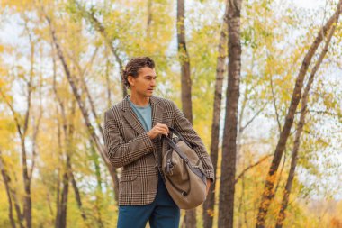 Kahverengi ceketli uzun boylu yakışıklı bir adam sonbahar sokağında çantasıyla yürüyor.