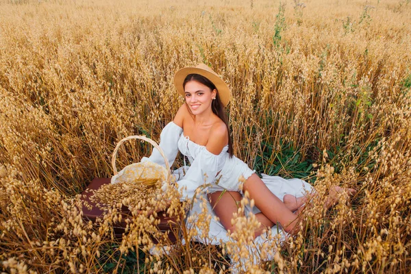 Kırsal Kır Sahnesi. Uzun saçlı, beyaz bluz ve hasır şapkalı güzel bir kadın altın yulaf tarlasında, sepetin yanında, yulaf kulaklı oturuyor..
