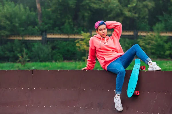 Portrait of a trendy pretty young girl with short colored hair and nose piercing dressed in pink hoodie and jeans sitting next to the skateboard court with her blue plastic skateboard. Copy space