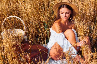 Kırsal Kır Sahnesi. Uzun saçlı, beyaz bluz ve hasır şapkalı güzel bir kadın altın yulaf tarlasında, sepetin yanında, yulaf kulaklı oturuyor..