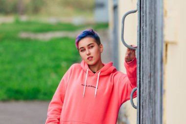 Kısa renkli saçları ve burnu olan pembe kapüşonlu piercingli genç kız ahşap kapılı duvarın yanında satanist gibi duruyor. Boşluğu kopyala.