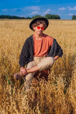 Kırsal Kır Sahnesi. Siyah gömlekli, siyah şapkalı ve pembe gözlükle altın yulaf tarlasında kahverengi deri bir valizde oturan uzun boylu yakışıklı bir adam. Mavi gökyüzü ile yaz manzarası