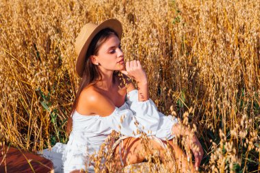 Kırsal Kır Sahnesi. Uzun saçlı, beyaz bluz ve hasır şapkalı güzel bir kadın altın yulaf tarlasında, sepetin yanında, yulaf kulaklı oturuyor..