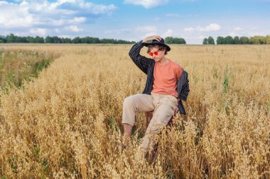 Kırsal Kır Sahnesi. Siyah gömlekli, siyah şapkalı ve pembe gözlükle altın yulaf tarlasında kahverengi deri bir valizde oturan uzun boylu yakışıklı bir adam. Mavi gökyüzü ile yaz manzarası
