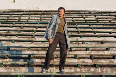 Gün batımında eski stadyumun ezilmiş tribününde kot ceket giymiş uzun boylu yakışıklı bir adamın moda portresi.