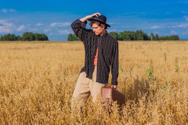 Kırsal Kır Sahnesi. Siyah gömlekli ve siyah şapkalı uzun boylu yakışıklı adam altın yulaf tarlasında kahverengi deri valiziyle dikiliyordu. Mavi gökyüzü ile yaz manzarası