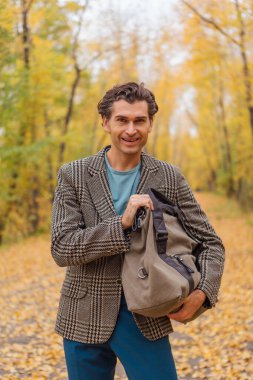 Kahverengi ceketli uzun boylu yakışıklı bir adam sonbahar sokağında çantasıyla yürüyor.