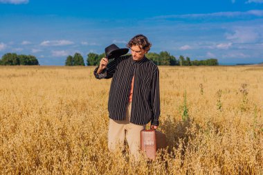 Kırsal Kır Sahnesi. Siyah gömlekli ve siyah şapkalı uzun boylu yakışıklı adam altın yulaf tarlasında kahverengi deri valiziyle dikiliyordu. Mavi gökyüzü ile yaz manzarası