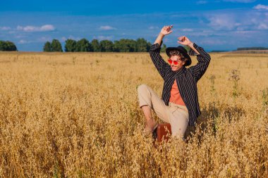 Kırsal Kır Sahnesi. Siyah gömlekli, siyah şapkalı ve pembe gözlükle altın yulaf tarlasında kahverengi deri bir valizde oturan uzun boylu yakışıklı bir adam. Mavi gökyüzü ile yaz manzarası