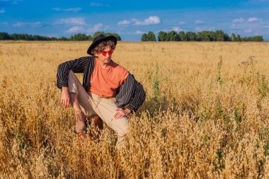 Kırsal Kır Sahnesi. Siyah gömlekli, siyah şapkalı ve pembe gözlükle altın yulaf tarlasında kahverengi deri bir valizde oturan uzun boylu yakışıklı bir adam. Mavi gökyüzü ile yaz manzarası