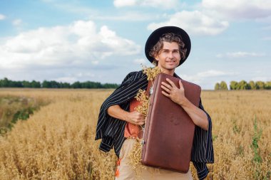 Kırsal Kır Sahnesi. Siyah gömlekli ve siyah şapkalı uzun boylu yakışıklı adam altın yulaf tarlasında kulaklarla dolu kahverengi klasik deri bavula sarılıyor.