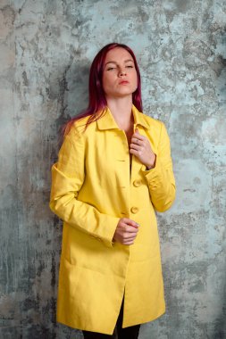 Sarı yağmurluk giymiş güzel bir kadın yüzüne dokunuyor ve beton duvarın yanında poz veriyor..