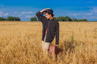 Kırsal Kır Sahnesi. Siyah gömlekli ve siyah şapkalı uzun boylu yakışıklı adam altın yulaf tarlasında kahverengi deri valiziyle dikiliyordu. Mavi gökyüzü ile yaz manzarası