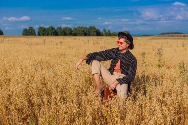 Kırsal Kır Sahnesi. Siyah gömlekli, siyah şapkalı ve pembe gözlükle altın yulaf tarlasında kahverengi deri bir valizde oturan uzun boylu yakışıklı bir adam. Mavi gökyüzü ile yaz manzarası