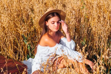 Kırsal Kır Sahnesi. Uzun saçlı, beyaz bluz ve hasır şapkalı güzel bir kadın altın yulaf tarlasında, sepetin yanında, yulaf kulaklı oturuyor..