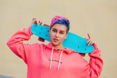 Omuzlarında mavi plastik kaykay tutan kısa saçlı şık genç bir kadın. Gençlik kavramı.