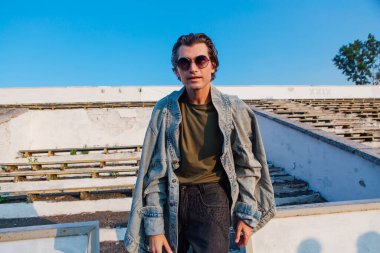 Uzun boylu, kot ceket giymiş yakışıklı bir adamın moda portresi güneş gözlüğü eski stadyumun ezilmiş tribününde gün batımında.