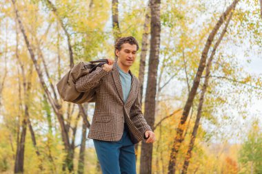 Kahverengi ceketli uzun boylu yakışıklı bir adam sonbahar sokağında çantasıyla yürüyor.
