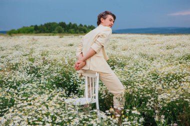 Beyaz takım elbiseli uzun boylu, yakışıklı bir adam papatya çiçekleri tarlasında bir sandalyenin arkasında oturuyordu.