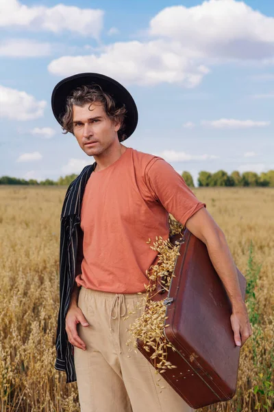 Tall handsome man holding brown suitcase full of ears of oats at golden oat field. — Stock Photo, Image