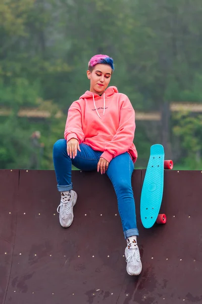 Portrait of a trendy pretty young gir with short colored hair sitting next to the skateboard court with her plastic skateboard. — Stock Photo, Image