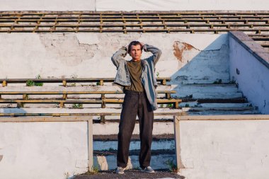 Gün batımında eski stadyumun ezilmiş tribününde kot ceket giymiş uzun boylu yakışıklı bir adamın moda portresi.
