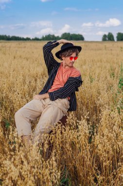 Kırsal Kır Sahnesi. Siyah gömlekli, siyah şapkalı ve pembe gözlükle altın yulaf tarlasında kahverengi deri bir valizde oturan uzun boylu yakışıklı bir adam. Mavi gökyüzü ile yaz manzarası