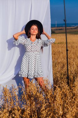 Kırsal Kır Sahnesi. Beyaz benekli elbiseli, siyah şapkalı, uzun saçlı, altın yulaf tarlasında beyaz perdeli genç bir kadın..