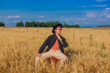 Kırsal Kır Sahnesi. Siyah gömlekli, siyah şapkalı ve pembe gözlükle altın yulaf tarlasında kahverengi deri bir valizde oturan uzun boylu yakışıklı bir adam. Mavi gökyüzü ile yaz manzarası
