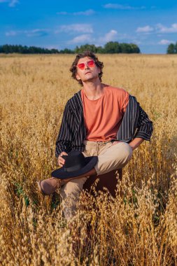 Kırsal Kır Sahnesi. Siyah gömlekli, siyah şapkalı ve pembe gözlükle altın yulaf tarlasında kahverengi deri bir valizde oturan uzun boylu yakışıklı bir adam. Mavi gökyüzü ile yaz manzarası