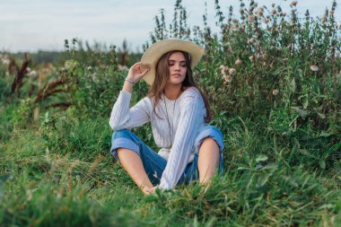 Beyaz kazak, kot pantolon ve kovboy hasır şapkası giyen genç, güzel esmer kadın yeşil çimlerin üzerinde oturuyor, gün batımında gülümsüyor ve gülüyor.