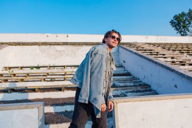 Uzun boylu, kot ceket giymiş yakışıklı bir adamın moda portresi güneş gözlüğü eski stadyumun ezilmiş tribününde gün batımında.