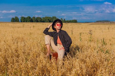 Kırsal Kır Sahnesi. Siyah gömlekli, siyah şapkalı ve pembe gözlükle altın yulaf tarlasında kahverengi deri bir valizde oturan uzun boylu yakışıklı bir adam. Mavi gökyüzü ile yaz manzarası