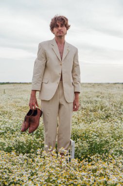 Uzun boylu, beyaz takım elbiseli, çıplak vücutlu, papatya çiçekli sandalyede çıplak ayakla duran ve elinde ayakkabıları olan yakışıklı bir adam.